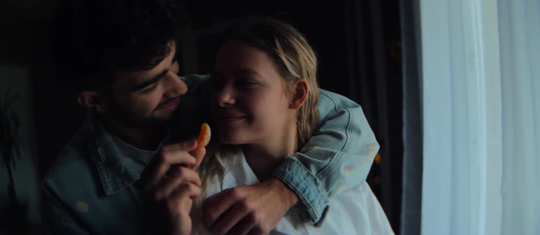 A loving man embraces his girlfriend from behind as she sips her drink by the window, playfully feeding her a slice of orange