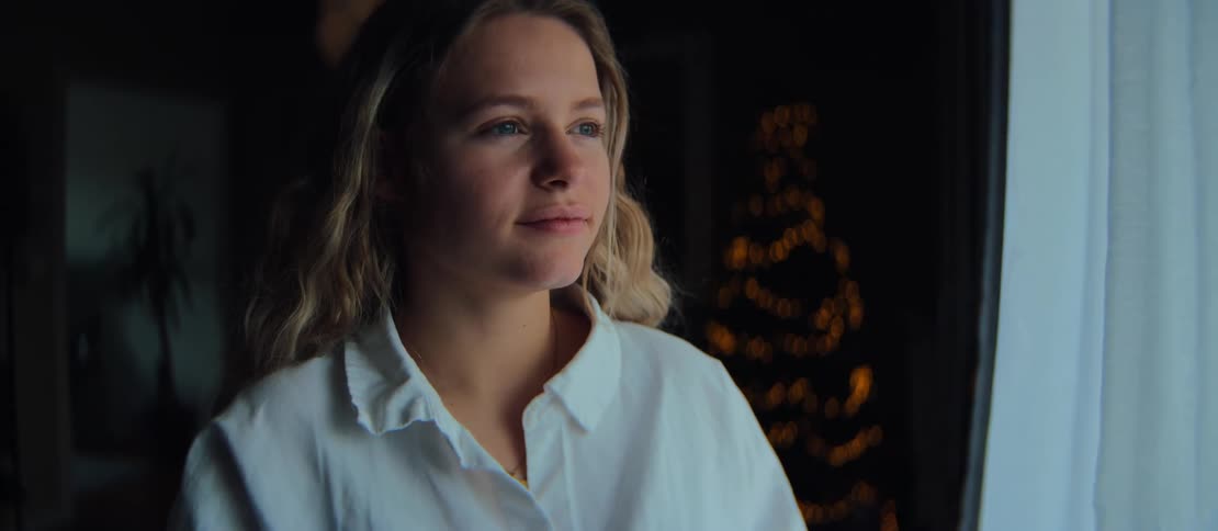 A contemplative young woman sips a warm beverage from a mug while gazing out a window, with a softly glowing Christmas tree in the background