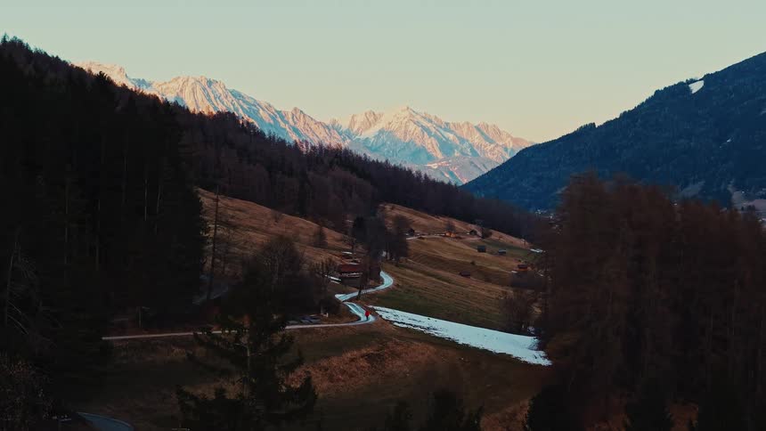 A solitary person in a red jacket hikes along a winding road through a peaceful mountain valley with majestic snow-capped peaks in the distance