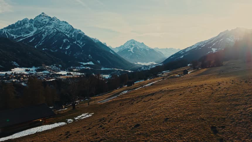 A breathtaking aerial shot flies over a golden hillside towards a majestic, snow-capped mountain valley at sunset