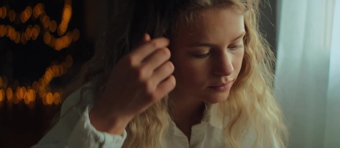 A close-up of a pensive young woman with curly blonde hair, focused and looking down in a softly lit room
