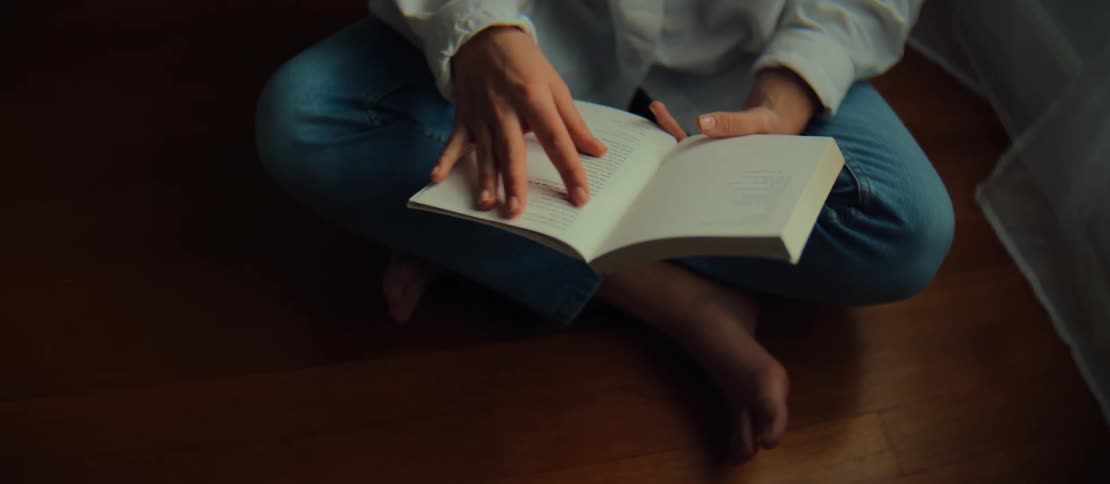A person sits cross-legged and barefoot on a wooden floor, slowly flipping the pages of a book in a calm setting