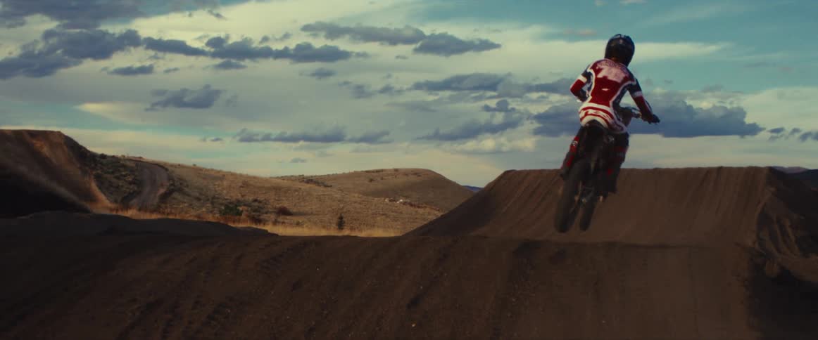 A motocross rider on a dirt track launches off a jump and kicks up a huge cloud of dust in dramatic slow motion