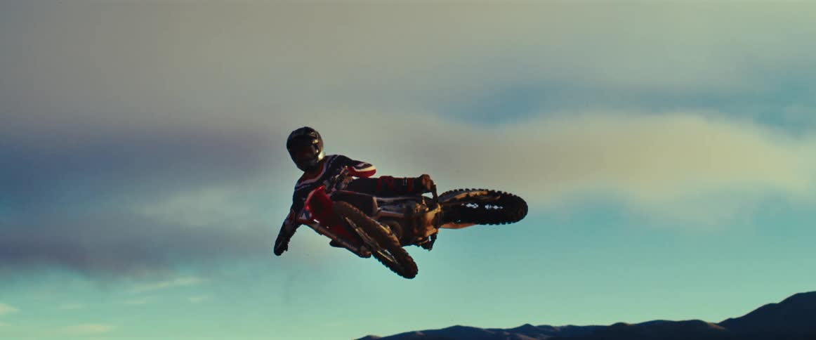 A freestyle motocross rider performs an impressive stunt, suspended in mid-air in slow motion with mountains in the background