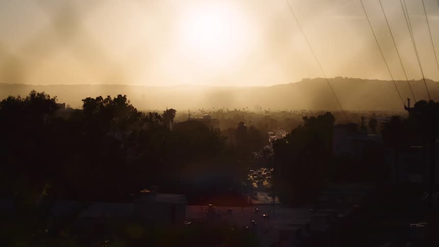 Slow dolly shot reveals a hazy, golden sunrise over a sprawling city valley, moving past silhouetted foliage and power lines