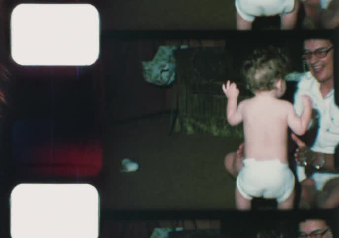 Vintage 8mm home movie captures a woman helping a curly-haired baby take their first steps in a living room