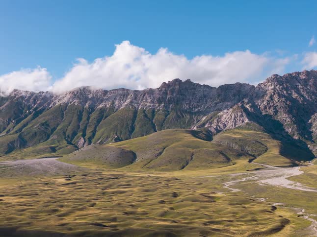 A time-lapse of clouds and shadows sweeping across a vast, green valley at the foot of a rugged mountain range