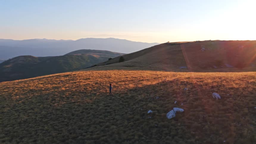An aerial shot follows a woman hiking across a vast, golden mountain landscape at sunrise, revealing the ancient Rocca Calascio castle ruins in Italy