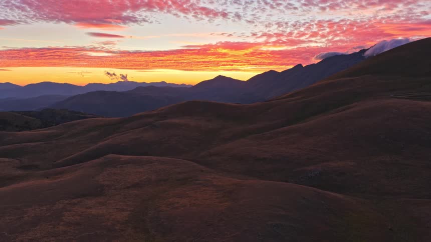 A breathtaking, fiery sunset with vibrant pink and orange clouds is revealed over a vast, rolling mountain landscape