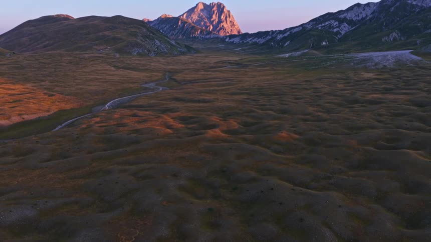 Epic aerial tilt-up reveals the majestic Gran Sasso mountain range bathed in the warm glow of sunrise over the vast Campo Imperatore plateau