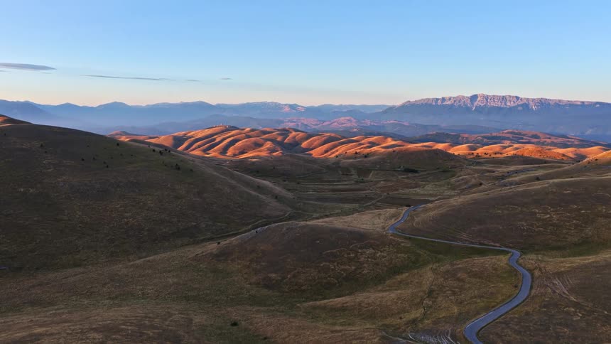 Aerial view of a scenic winding road through golden rolling hills at sunrise with mountains in the background