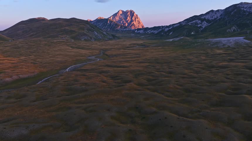 A slow tilt up reveals a majestic mountain range glowing in the warm light of sunset over a vast, tranquil valley
