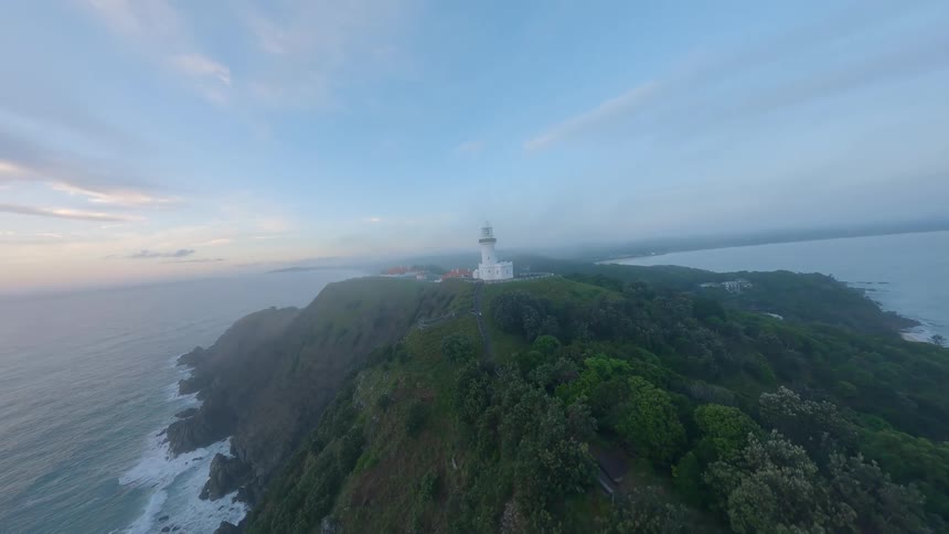 An ethereal aerial flight approaches the iconic Cape Byron Lighthouse as its beacon shines through the morning mist over the vast ocean