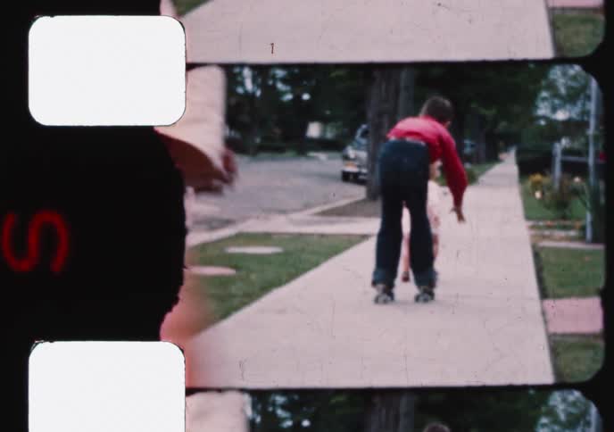 A toddler walks along a suburban sidewalk as two boys on roller skates follow behind in this vintage home movie clip