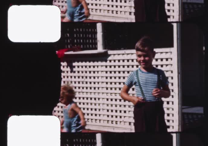 Vintage home movie from the 1950s shows a young boy in suspenders smiling at the camera while his little brother plays behind him