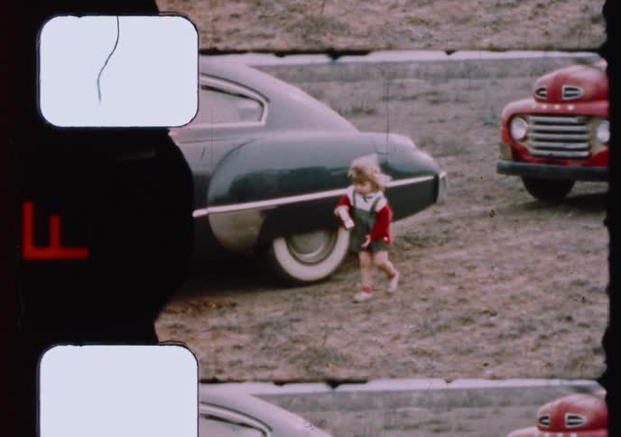 A curious little girl in a red sweater explores a dirt lot between two classic American cars in grainy, vintage 8mm footage