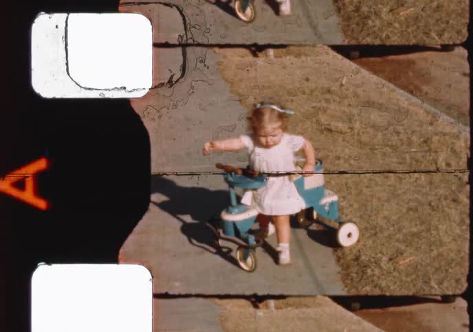 Archival 8mm film from the 1960s shows a toddler girl in a red coat and later a white dress, playing on her ride-on toys outdoors