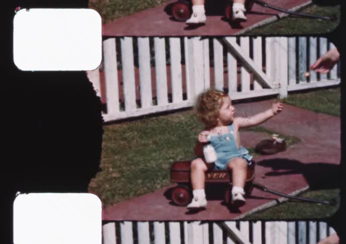 Authentic vintage footage shows a sweet toddler with curly blonde hair drinking milk from a bottle while sitting in a red wagon in the backyard