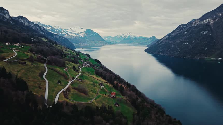 An epic aerial view captures the serene beauty of a vast lake surrounded by majestic mountains and rolling green hills under a cloudy sky