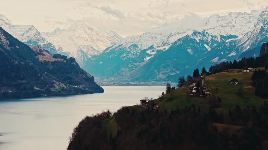 A majestic wide shot of a serene mountain lake in a valley, with snow-capped peaks in the background and a small village on a green slope