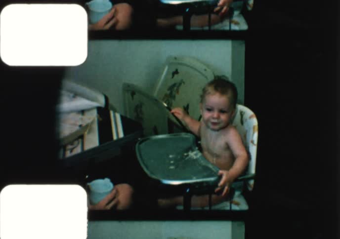 Vintage 8mm home movie captures a mother feeding her messy but happy baby boy in a high chair