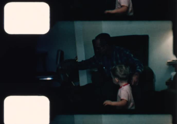 A candid 1970s home movie shows a happy toddler with a pacifier playing with a ball and his dog in the living room