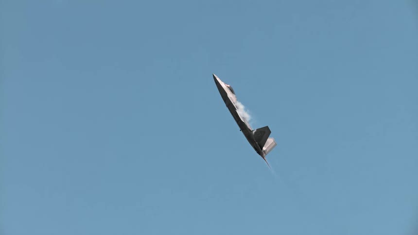 An F-22 Raptor fighter jet performs an impressive near-vertical climb in super slow motion against a clear blue sky