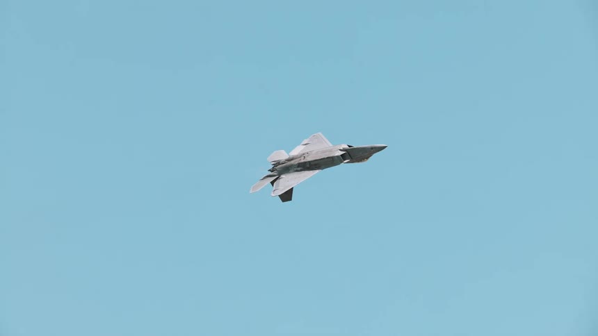An F-22 Raptor fighter jet performs a slow pass maneuver against a clear blue sky, showcasing its design and afterburners