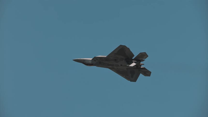 A sleek F-22 Raptor fighter jet executes a slow and graceful maneuver against a clear blue sky