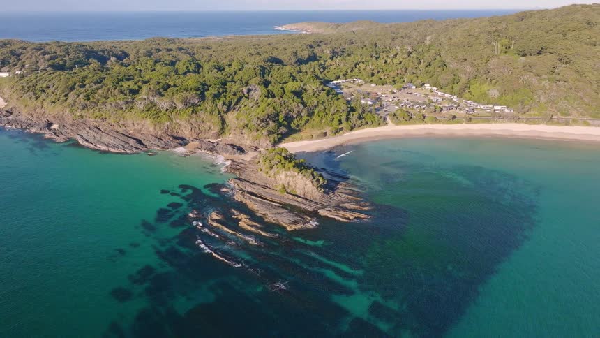 An aerial drone shot slowly pulls back to reveal a spectacular coastline featuring a rocky headland, clear turquoise water, dense forest, and a secluded beach campground