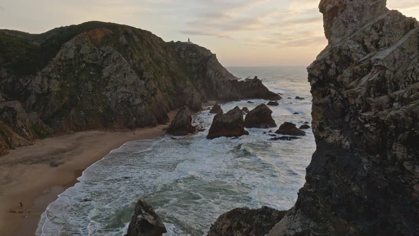 Majestic aerial reveal of a rugged coastline and a secluded beach as the sun sets over the ocean