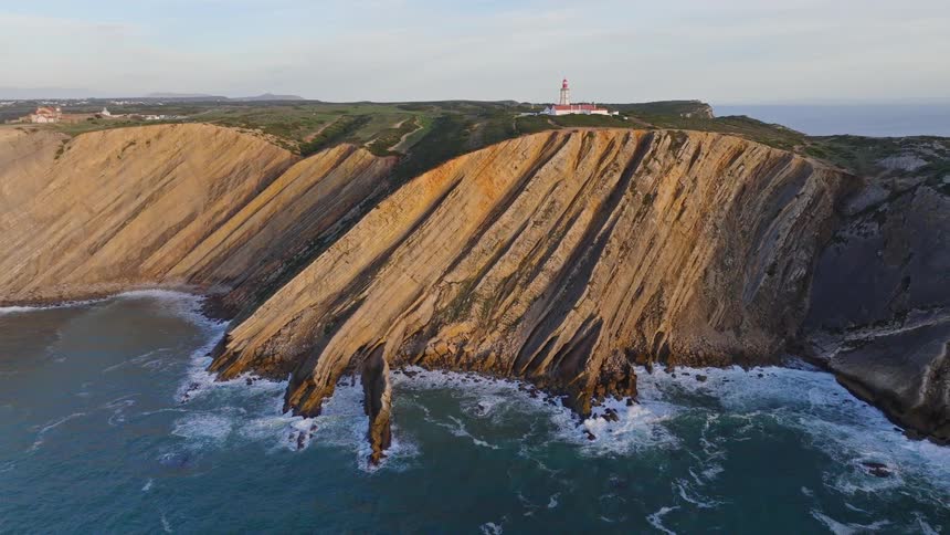 A stunning aerial view shows the dramatic, layered rock formations of a coastal cliff with a lighthouse perched on top as the ocean waves crash below