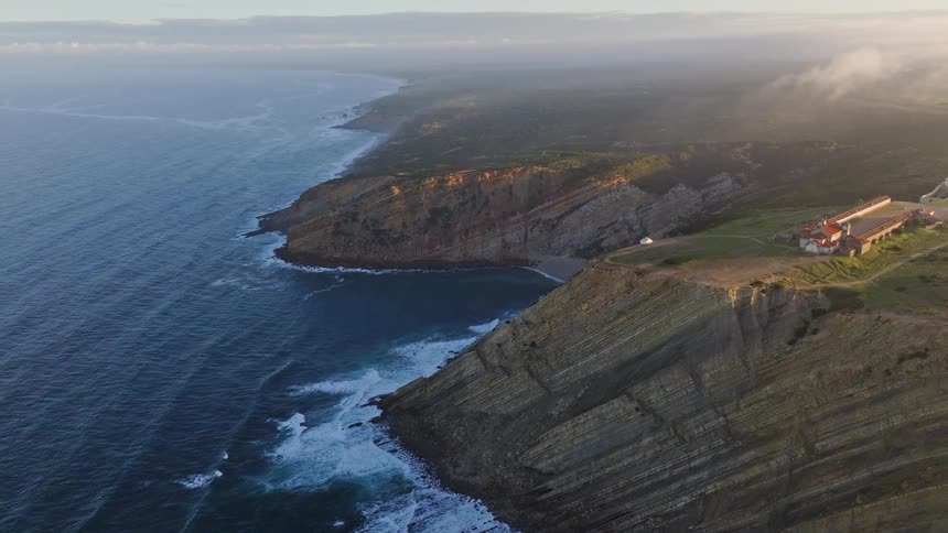 A serene aerial view shows the majestic layered cliffs of the Cabo Espichel coastline meeting the vast Atlantic Ocean at sunrise
