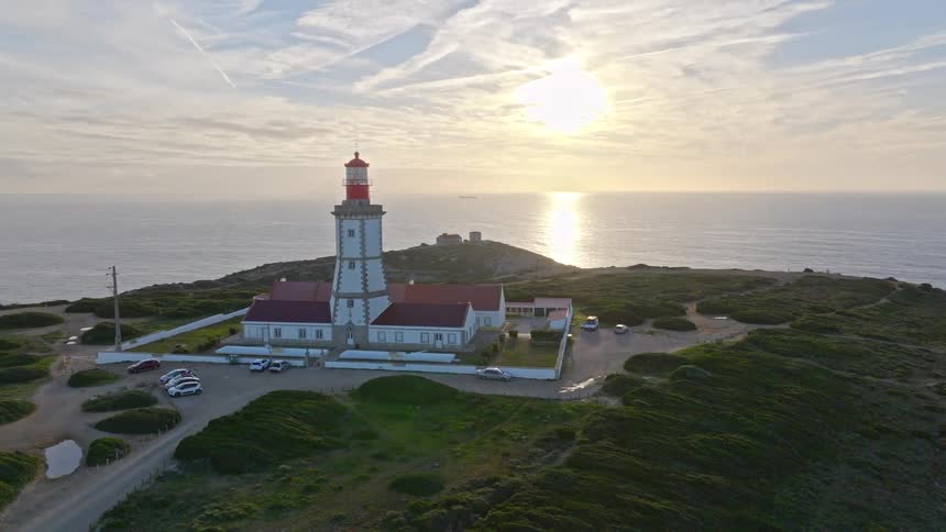 An aerial drone flies toward the historic Cabo Espichel Lighthouse on a verdant cliff overlooking the Atlantic Ocean as the sun sets on the horizon