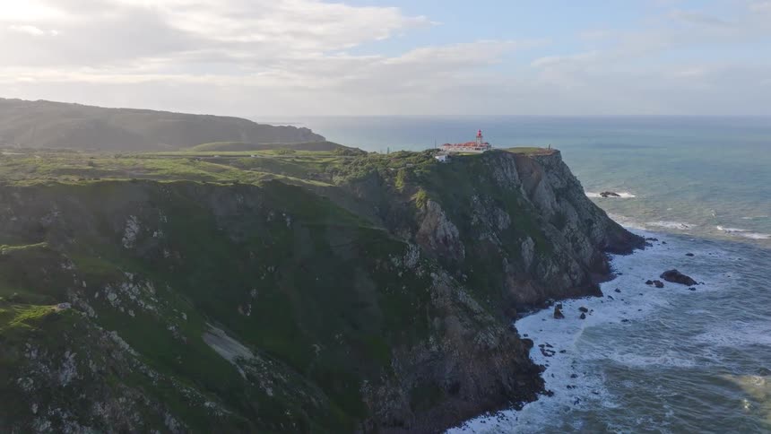 An epic aerial view slowly approaches the historic lighthouse at Cabo da Roca, Portugal, perched atop rugged green cliffs overlooking the vast Atlantic Ocean