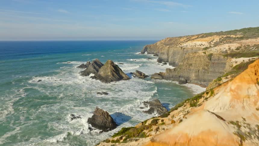 Aerial view of a person standing on the edge of a dramatic, colorful cliff, revealing a stunning, rugged coastline with waves crashing on the rocks below
