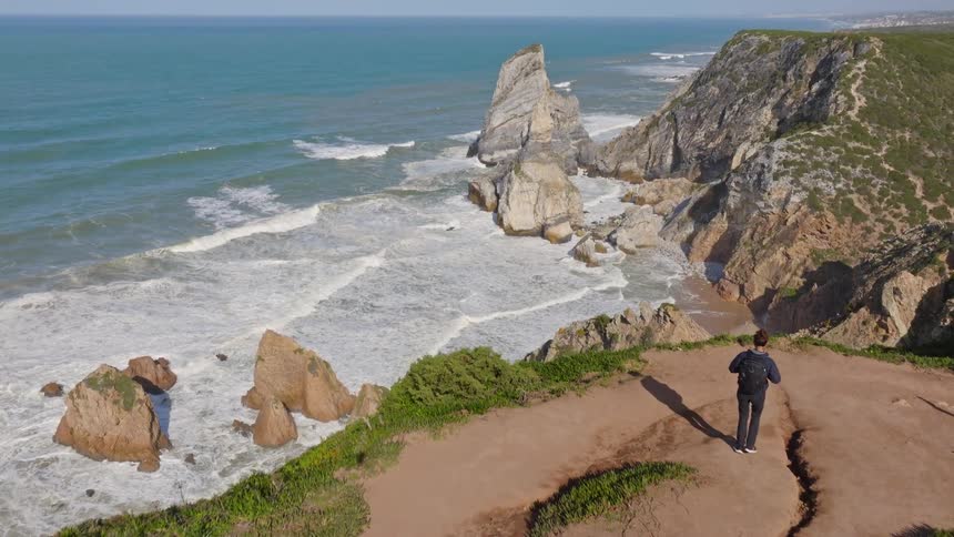 A hiker stands on a dramatic clifftop path, taking in the spectacular panoramic view of a rugged coastline and the vast ocean below