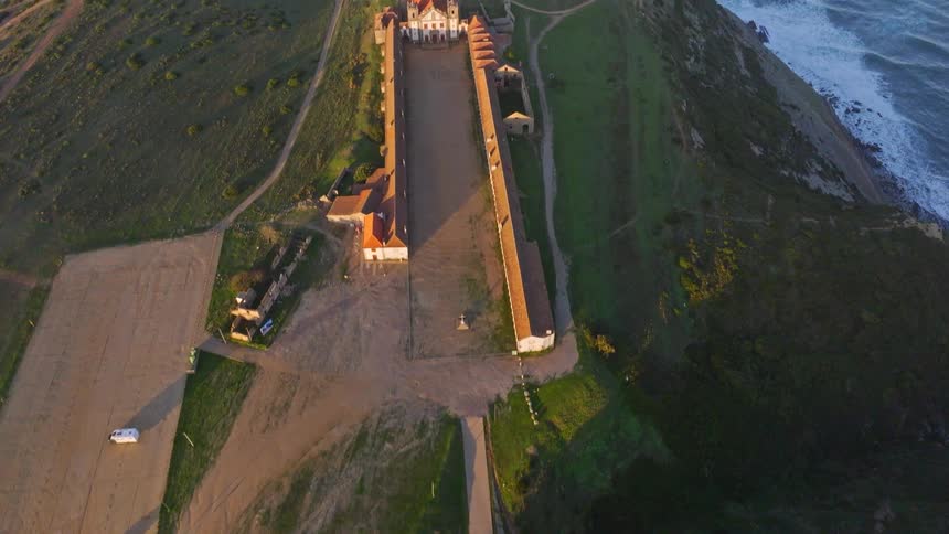 An aerial shot starts with a top-down view and slowly tilts up to reveal the Santuário de Nossa Senhora do Cabo Espichel in Portugal, perched on a cliff overlooking the ocean at sunset