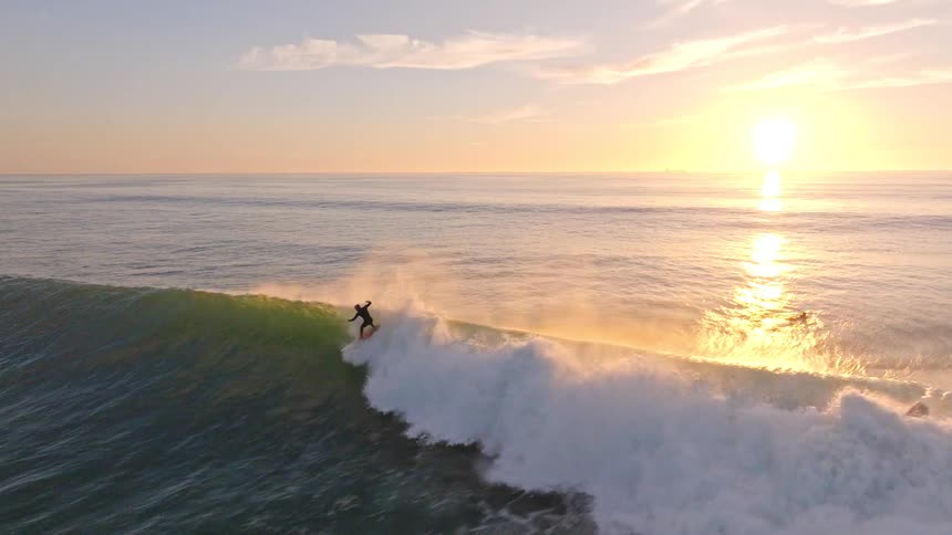 An aerial slow-motion shot captures a surfer skillfully riding a beautiful, sunlit wave during a golden sunset