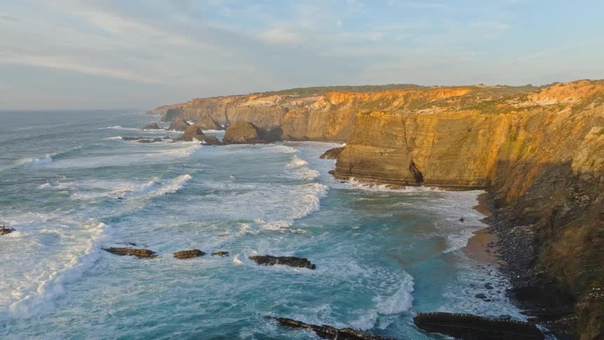 A beautiful aerial shot flies along a rugged, steep coastline where powerful ocean waves crash against the cliffs during a golden sunset