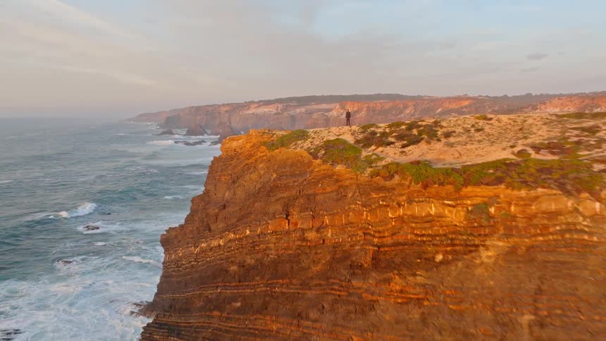 Inspiring aerial footage pushes in on a lone man standing atop a rugged coastal cliff watching the ocean waves at sunset