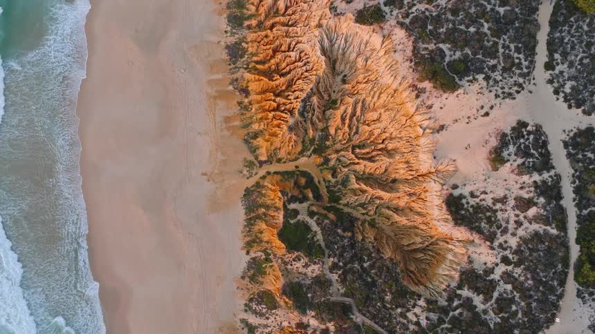 An awe-inspiring top-down aerial view slowly zooms out to reveal stunning eroded sandstone cliffs meeting the ocean waves on a sandy beach at golden hour
