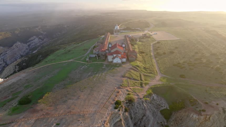 An aerial view begins with a top-down shot of the Santuario do Cabo Espichel, then dramatically pulls back to reveal the historic sanctuary perched on a rugged cliffside at sunset