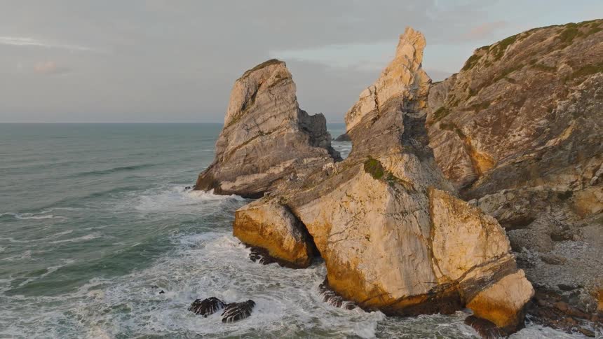 An aerial view pans around dramatic sea stacks and rugged cliffs as ocean waves crash against the shoreline during a beautiful sunset