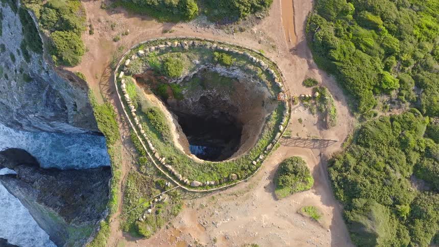 Top-down aerial view slowly ascends from a coastal sea cave, revealing a dramatic sinkhole on the clifftop next to the ocean