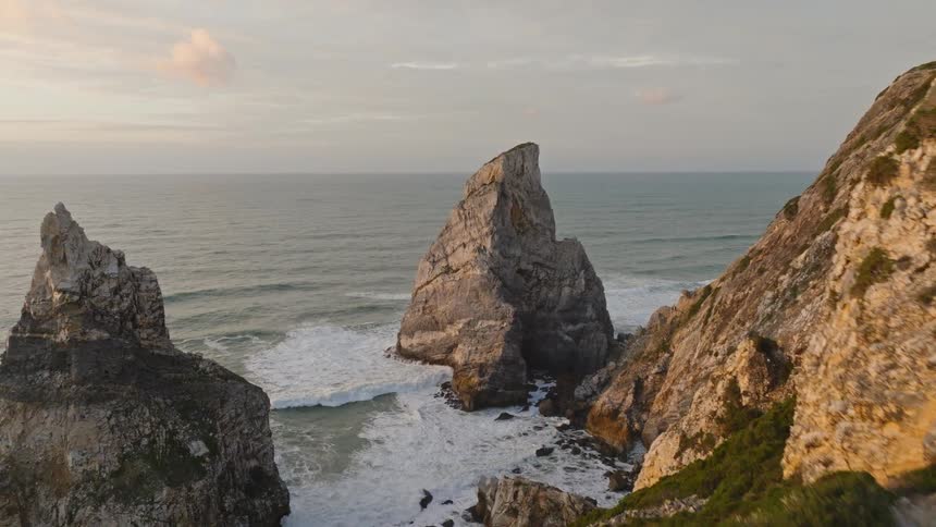 Aerial footage follows hikers along a winding path on a rugged, bush-covered cliffside, revealing majestic sea stacks in the ocean at sunset
