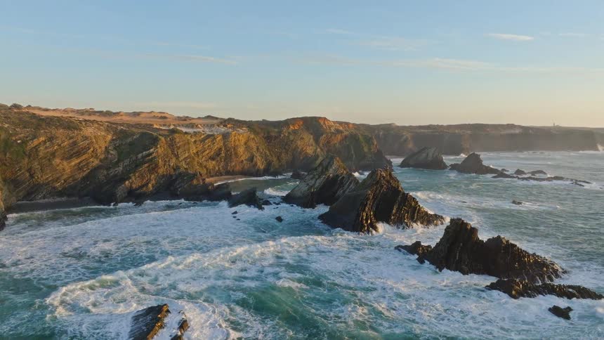Aerial footage captures the breathtaking beauty of a rugged coastline with dramatic rock formations and crashing ocean waves at golden hour