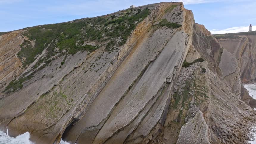 Aerial view pulls back from a stunningly detailed geological rock formation to reveal a massive coastal cliff with a lighthouse in the distance