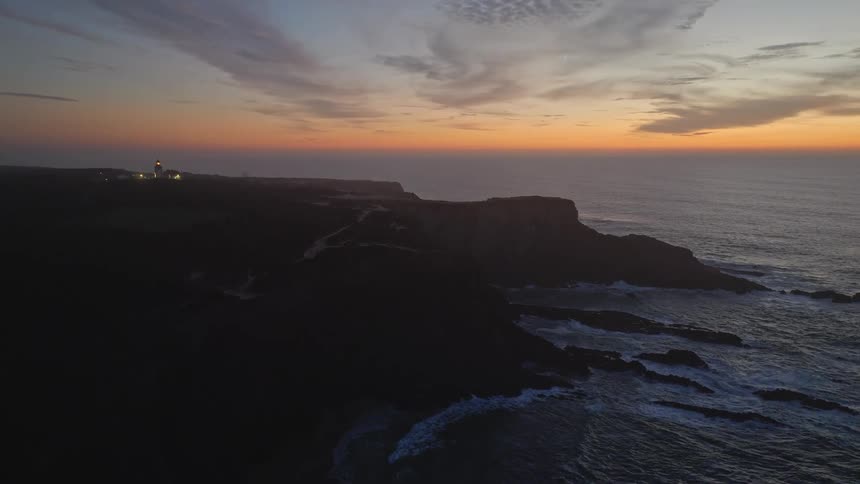 An aerial drone flies over a silhouetted coastline as a distant lighthouse flashes its light across the ocean at dusk