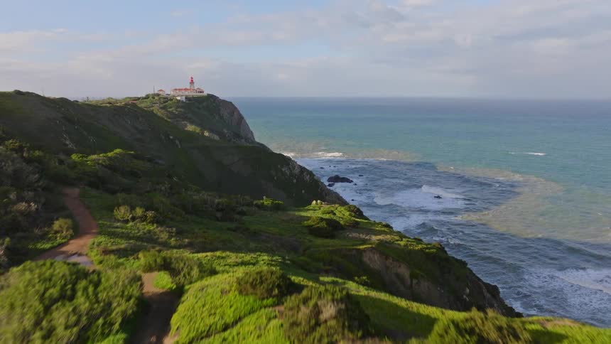 An aerial drone shot flies over vibrant green coastal hills to reveal a historic lighthouse perched atop a dramatic cliff overlooking the vast ocean